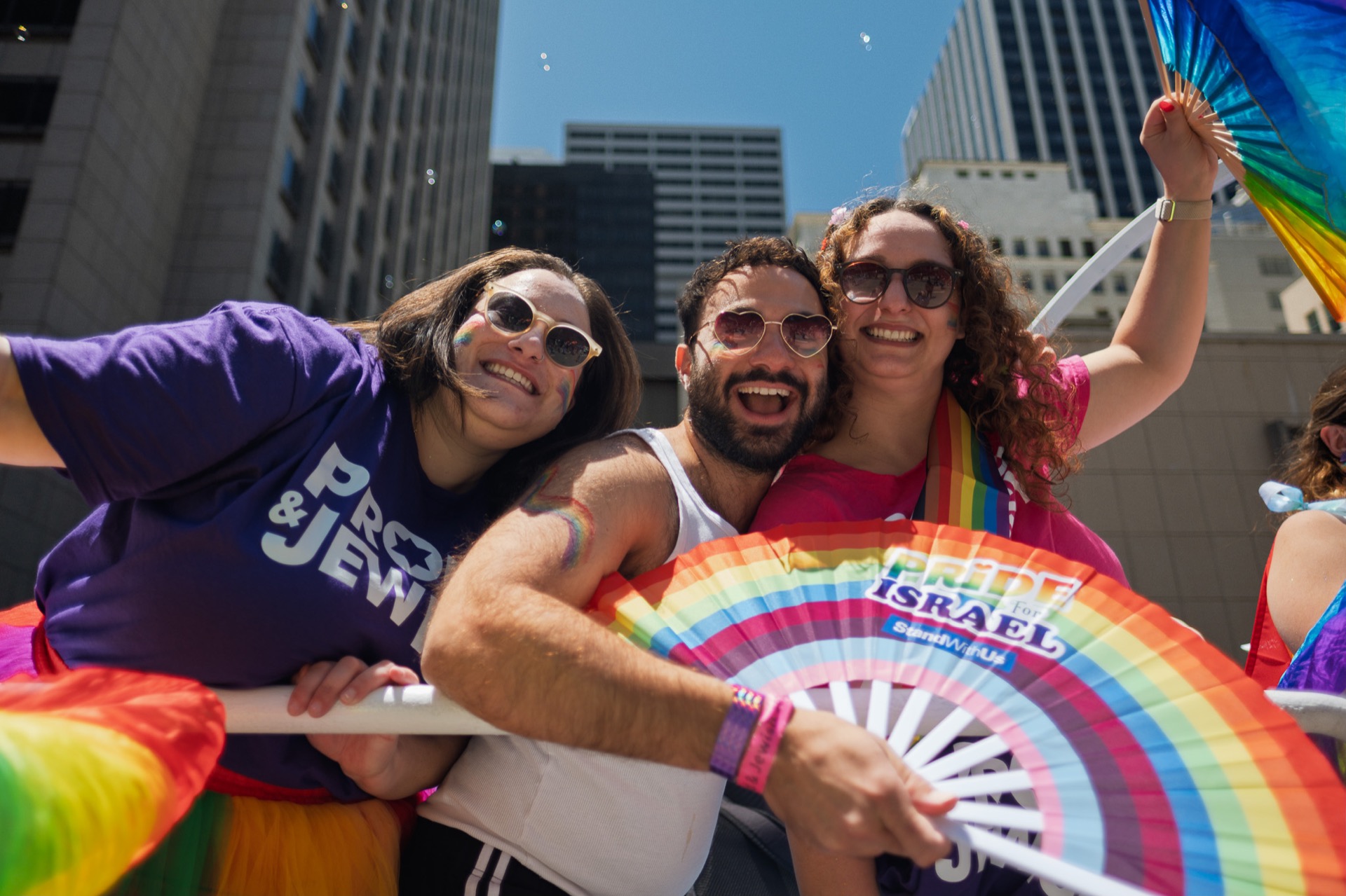 Three smiling people in rainbow clothing pose together with a rainbow fan at a Pride gathering.