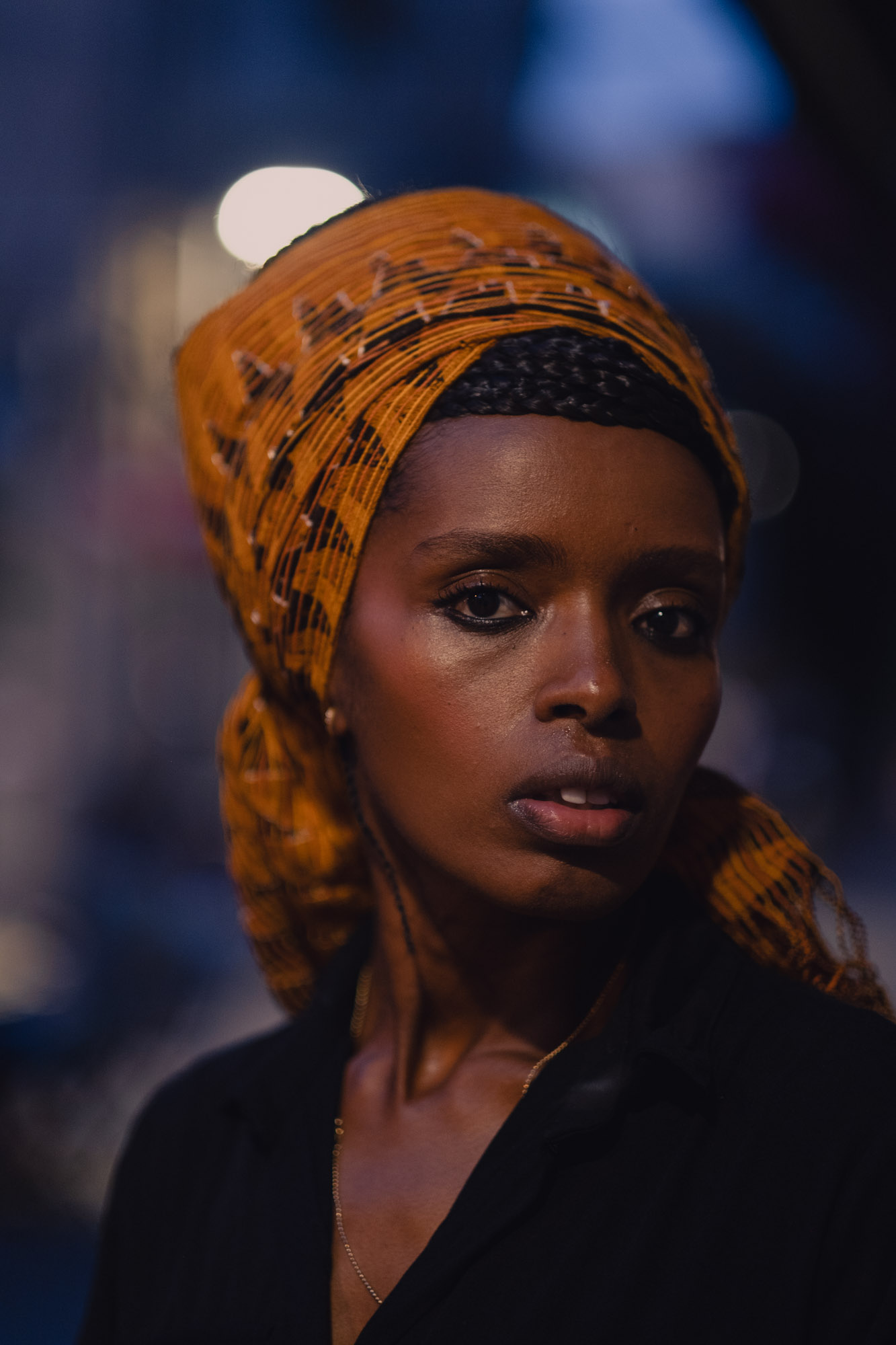A woman in an orange patterned headwrap looks toward the camera against a dark blue background.