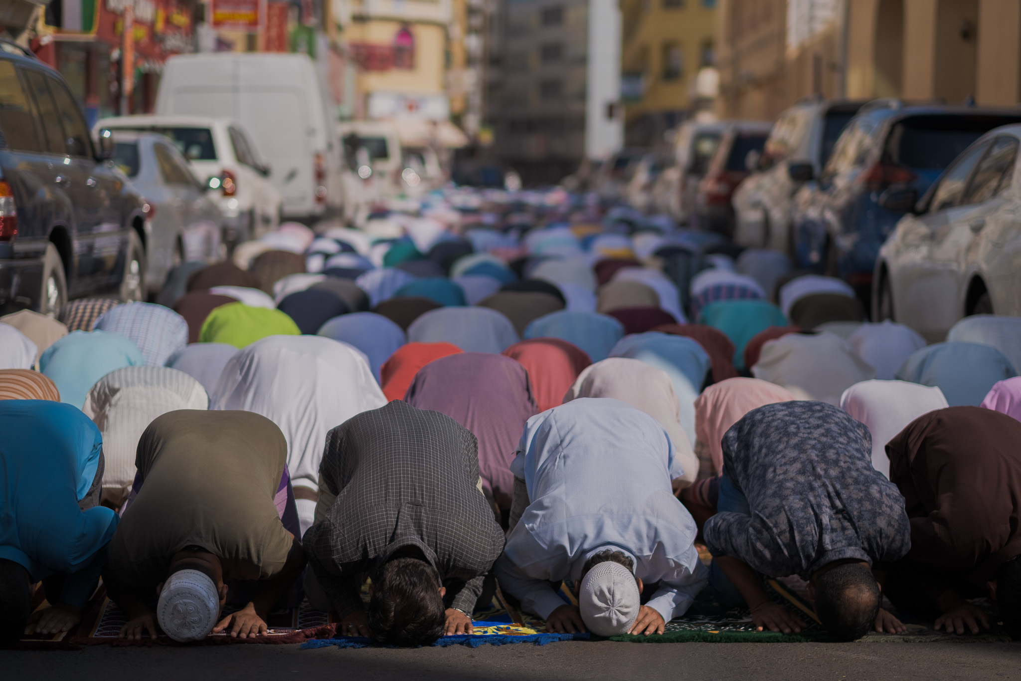 Rows of worshippers bow in prayer on rugs laid across a city street between parked cars.