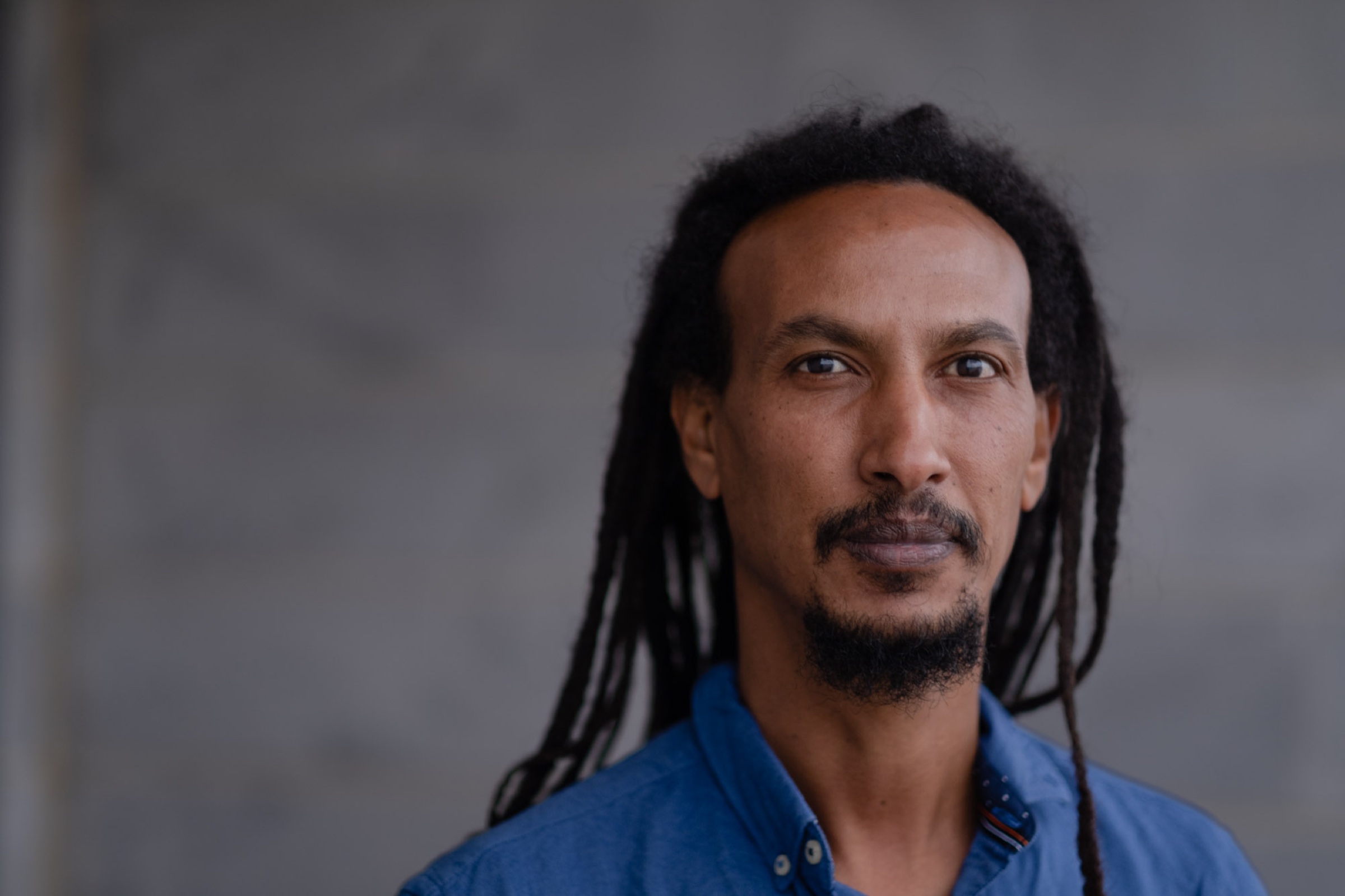 A man with long locs and a blue shirt looks beyond the camera against a neutral wall.