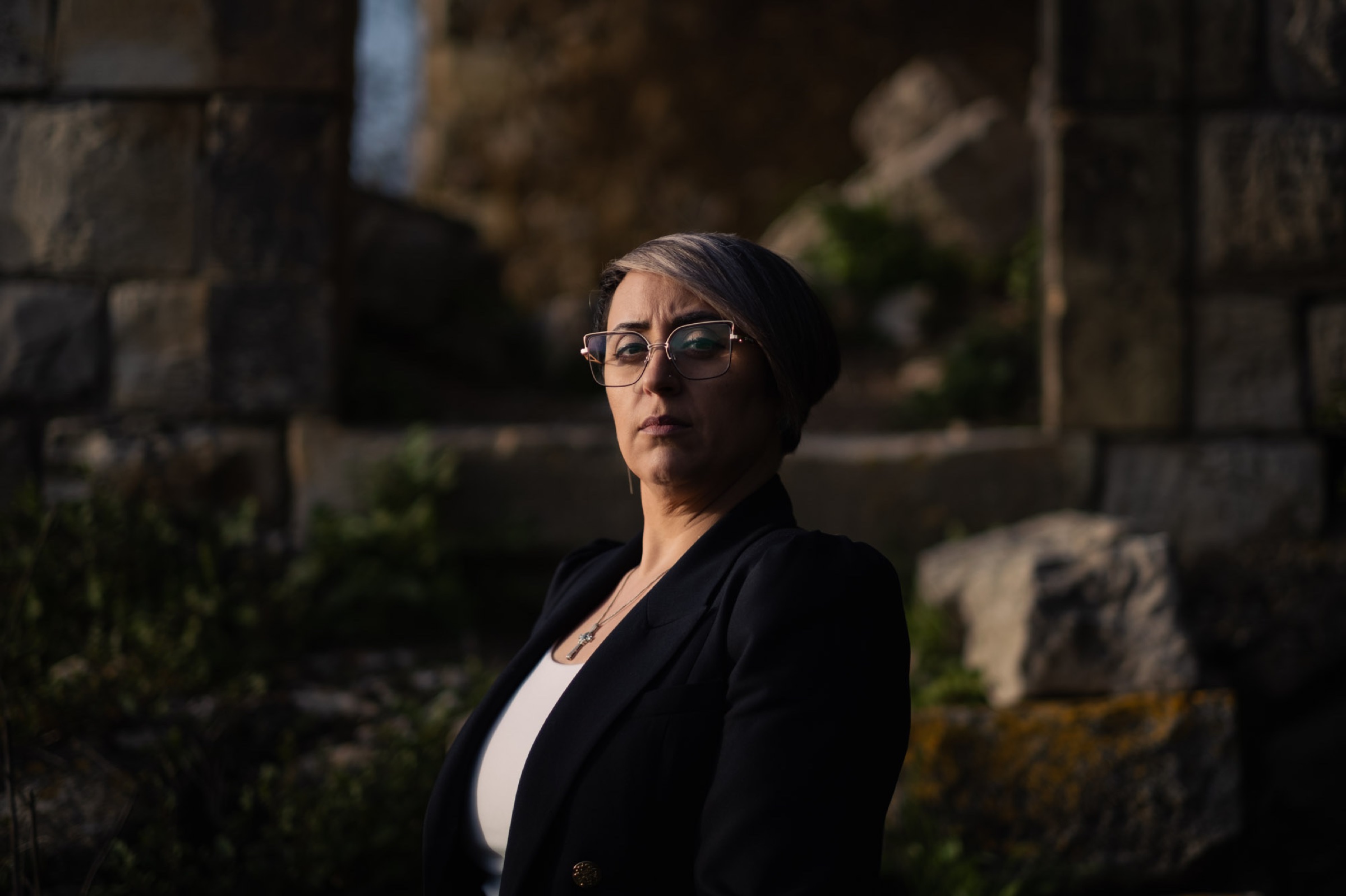 A woman wearing glasses and a dark blazer stands in directional light against monumental stone.
