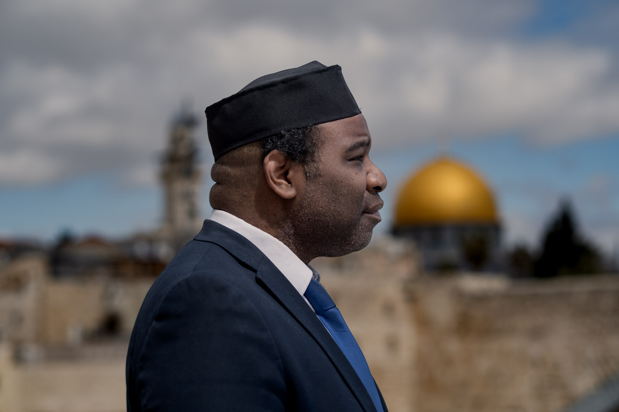 A man in a dark suit and cap is photographed in profile with the Dome of the Rock beyond him.