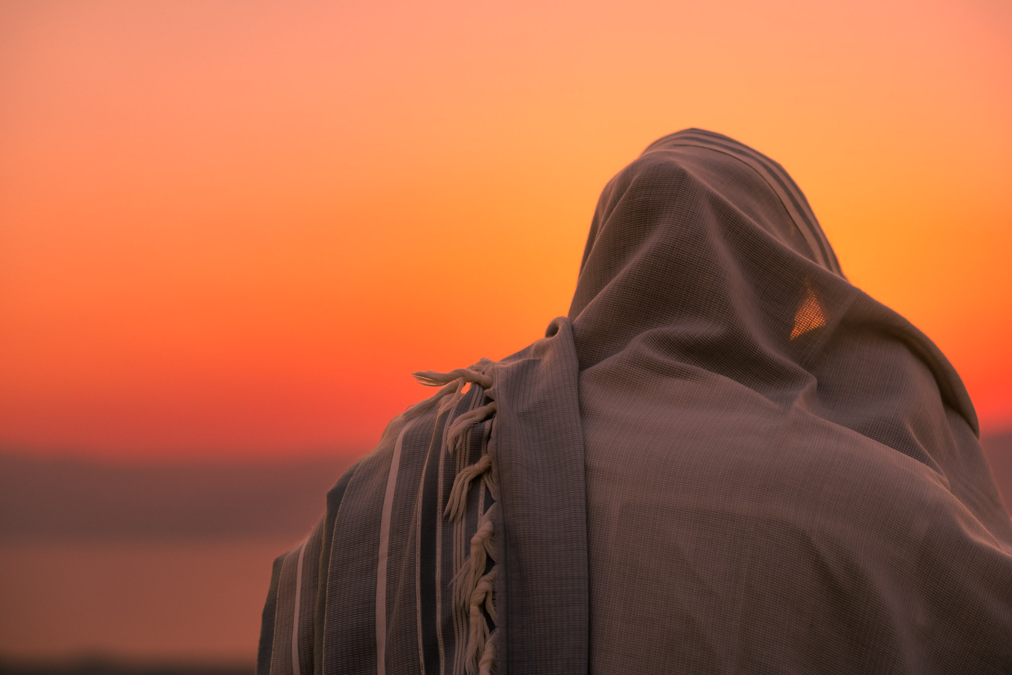 A person wrapped in a striped tallit stands against a vivid orange sunset.