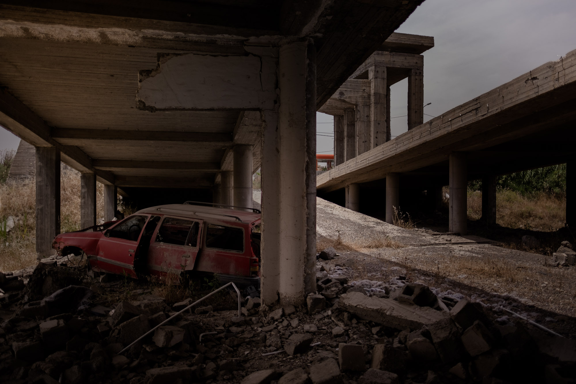 An abandoned red station wagon sits beneath unfinished concrete structures amid rubble.