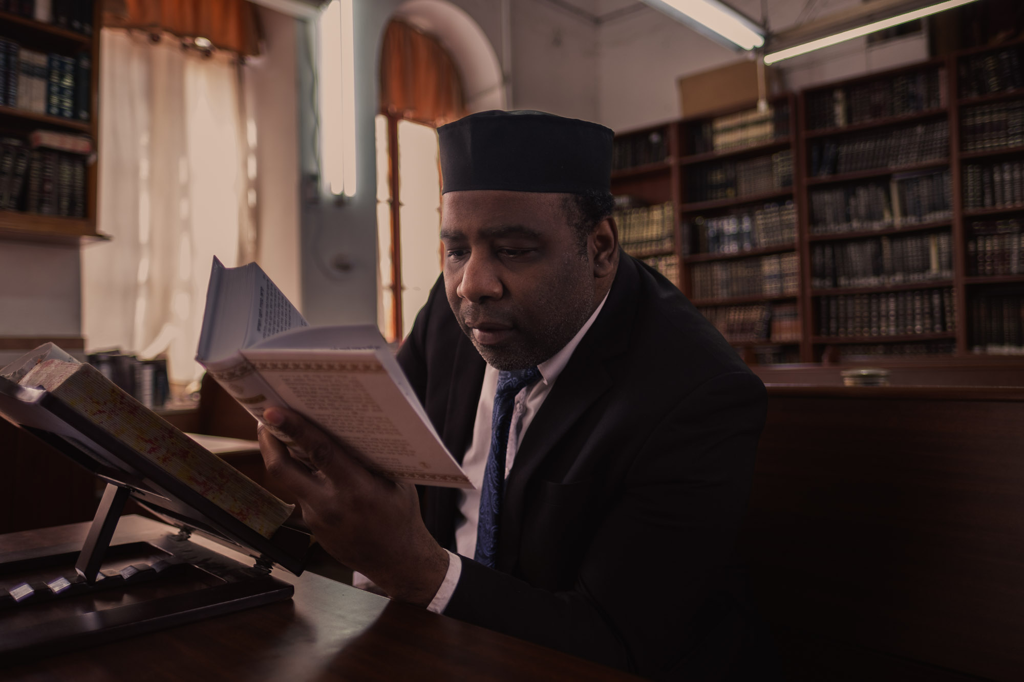 A man in a dark suit reads from an open book in a library lined with books.