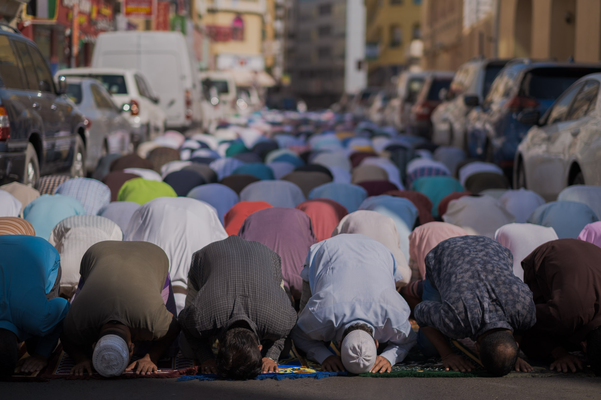 Rows of people bow in prayer along a city street between parked cars.